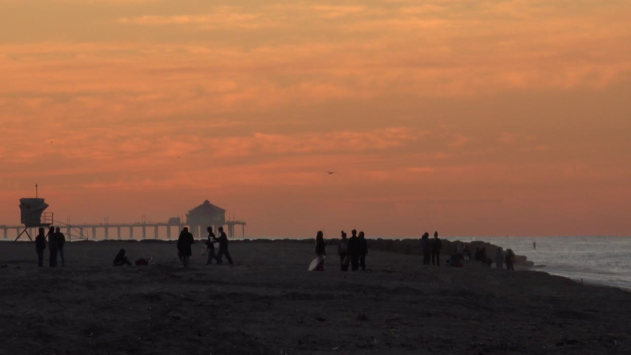 Silhouetted people stand and walk on the Huntington Beach shoreline at 0:00, with a lifeguard tower at left, pier structure in the distance, and waves along the right edge.