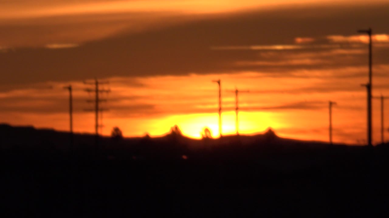 At 0:00 in Huntington Beach, the yellow sun sits just above a dark horizon with tree silhouettes, utility poles, streetlights, orange cloud bands, and no visible people.