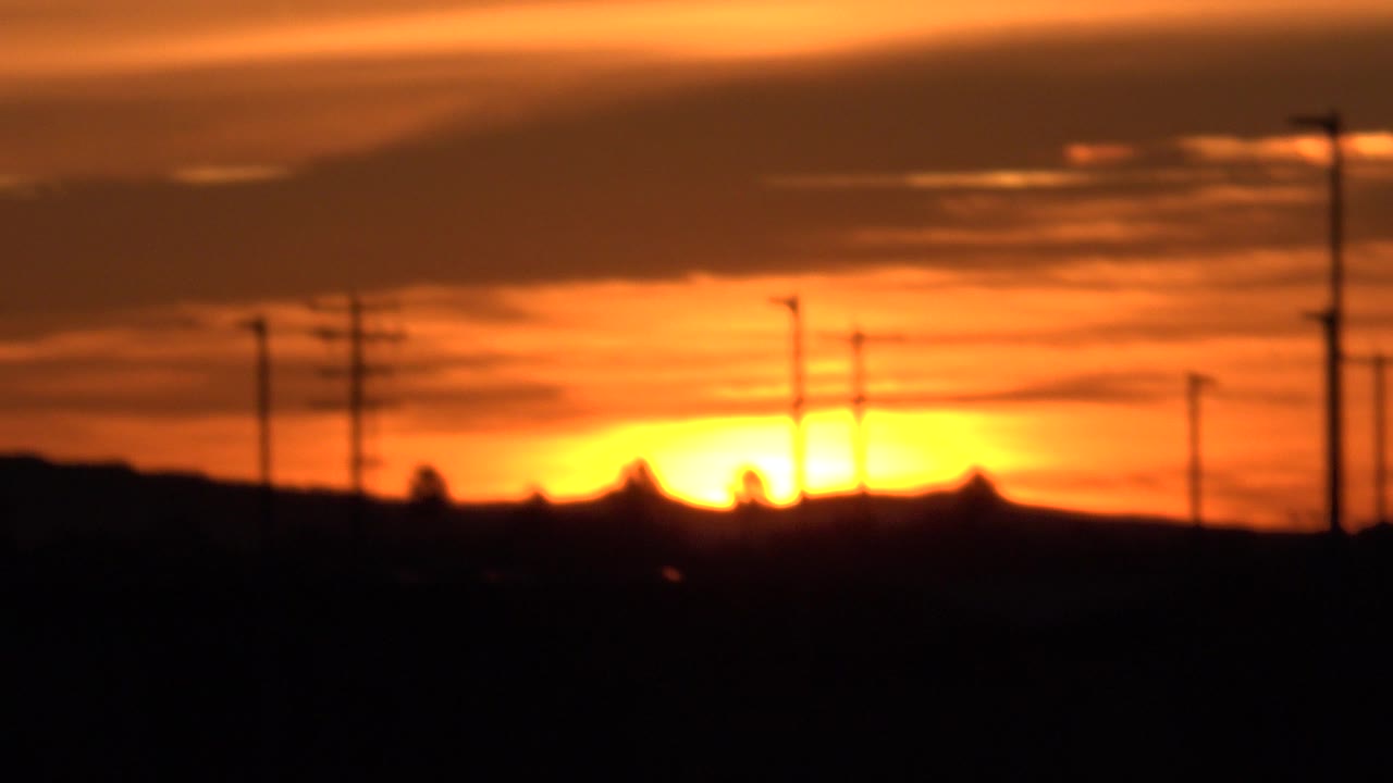 At 0:01 in Huntington Beach, the sun glows brighter near the center of the horizon while black pole and tree silhouettes cut into the orange sky, with no visible people.