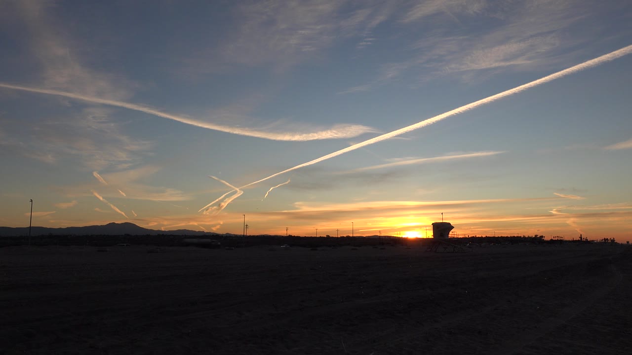Early frame of a Huntington Beach sunset with the sun low near the horizon, dark foreground land, distant hills, diagonal white cloud trails, and no visible people.