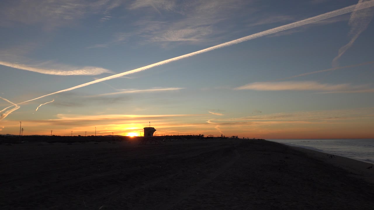 Huntington Beach sunset over a dark sandy beach with the ocean on the right, bright contrails overhead, a silhouetted tower near the horizon, and no visible people.