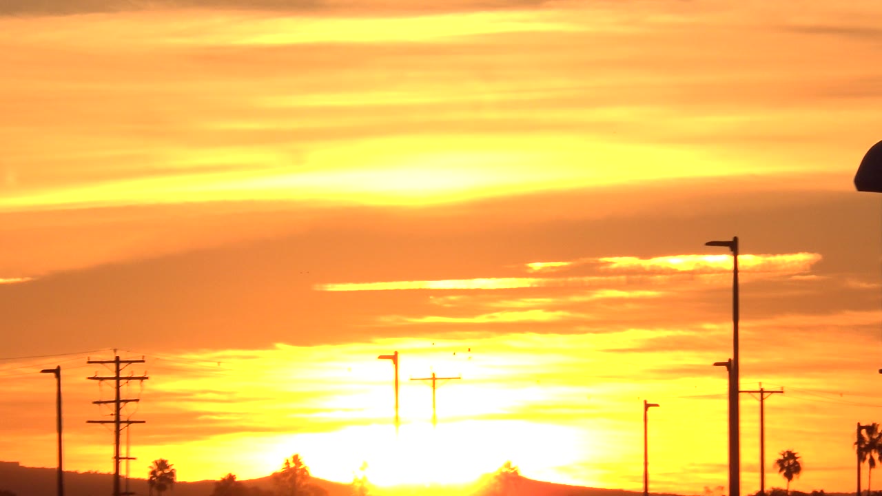 A bright orange Huntington Beach sunset sits low behind silhouetted streetlights, utility poles, distant landforms, and no visible people.
