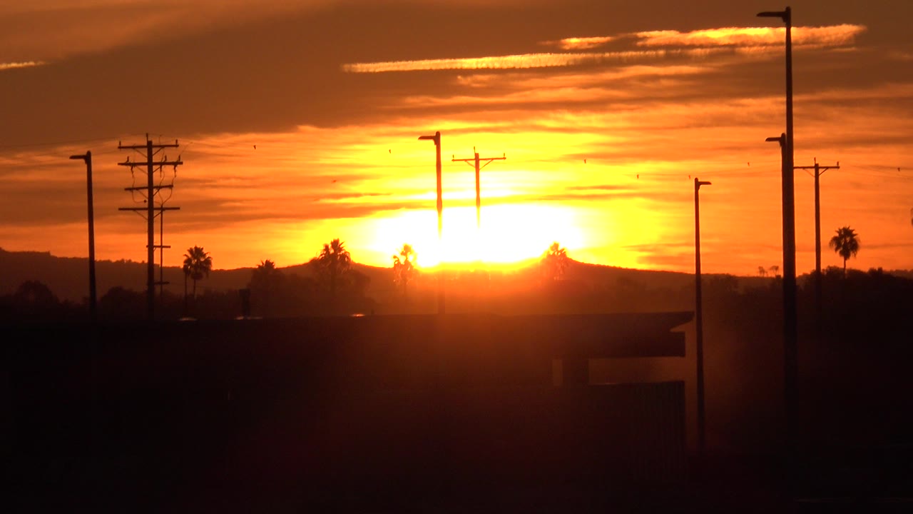 Opening frame of a Huntington Beach sunset with the sun low on the horizon, dark utility poles and streetlights in silhouette, orange clouds above, and no visible people.
