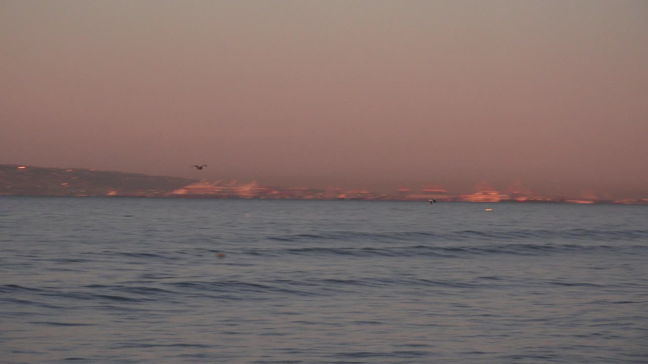 At Huntington Beach, California, low waves fill the foreground below a pink morning sky and distant coastal buildings, with no visible people.