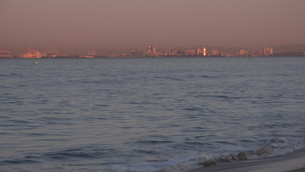 Huntington Beach, California view with rippled ocean in the foreground, a low wave near shore, distant buildings, and no visible people.