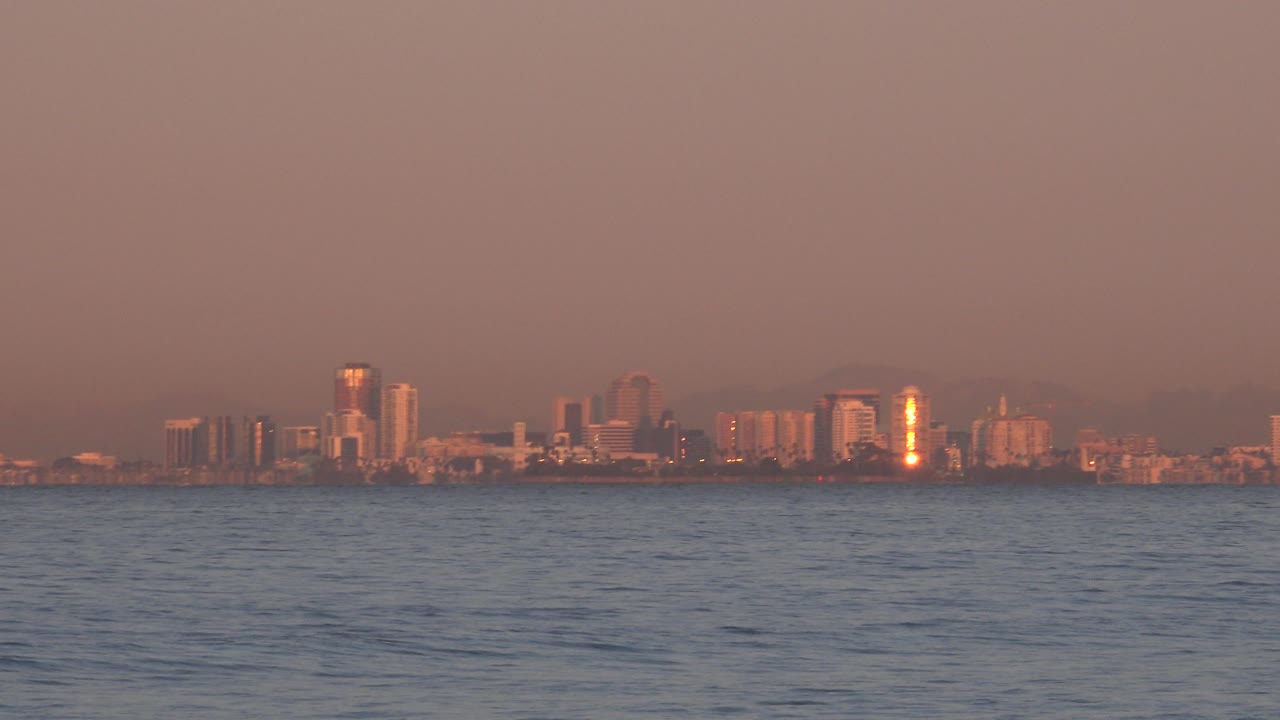 Huntington Beach, California appears as a low city silhouette beyond textured ocean water, with warm sunset haze and no visible people.