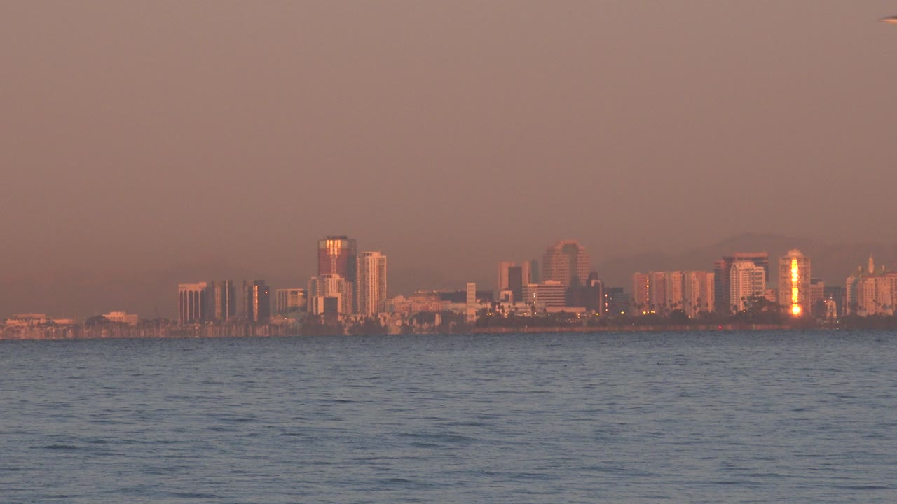 Huntington Beach skyline sits low on the horizon beyond blue ocean water under a hazy pink sunset sky, with no visible people.