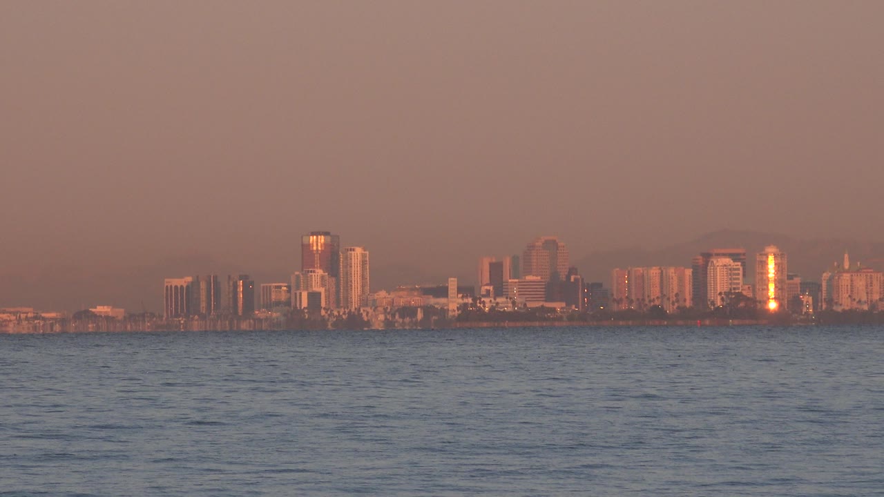 Distant shoreline buildings across ocean water under a hazy pink sky.