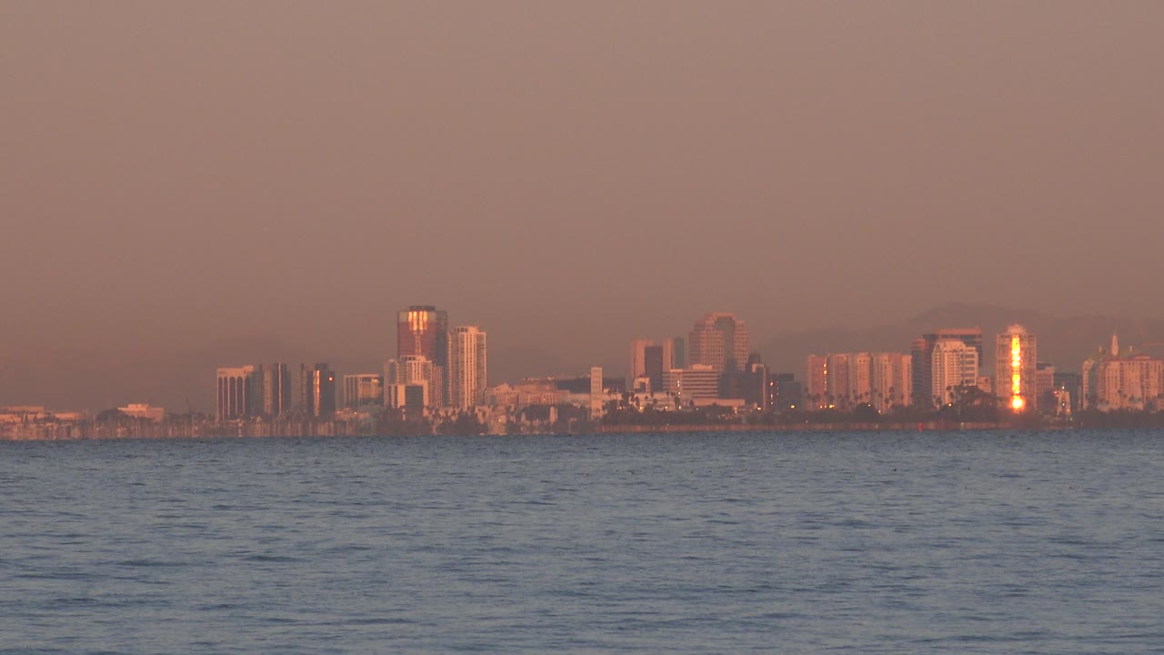 A wide view across the ocean shows distant Huntington Beach buildings, a bright vertical reflection on one tower, soft haze, and no visible people.