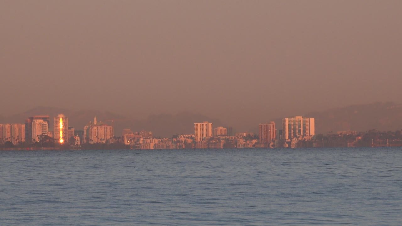 Huntington Beach buildings sit low on the horizon beyond rippled ocean water, with a bright vertical reflection on one tower and no visible people.
