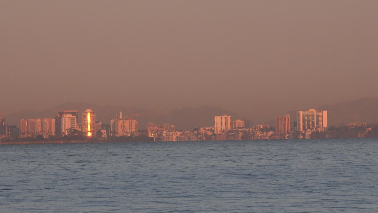 A wide Huntington Beach bay view shows low water in the foreground and distant city buildings under hazy orange sunset light, with no visible people.