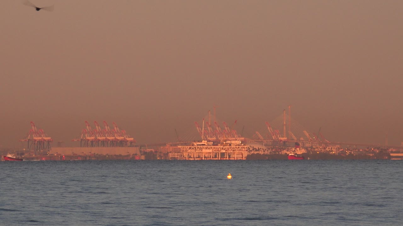 A bird flies high above the ocean at Huntington Beach, California, with distant pier and crane-like structures on the horizon in warm sunset haze and no visible people.