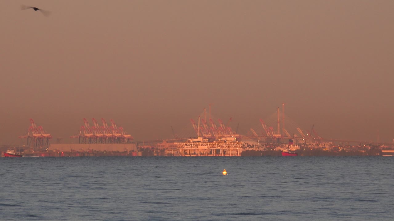 The Huntington Beach ocean fills the foreground while distant pier structures and cranes sit low on the horizon under orange sunset light, with no visible people.