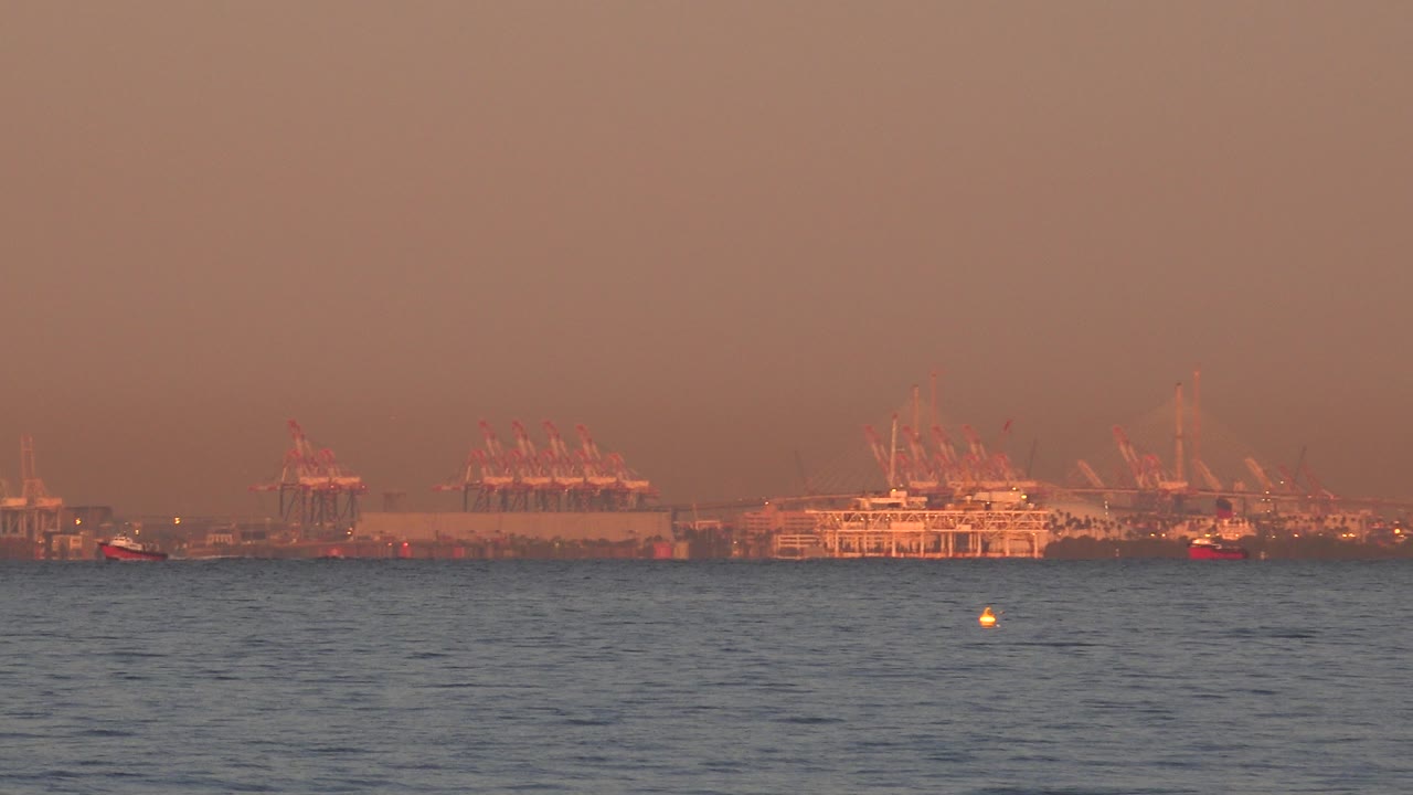 Huntington Beach ocean view at 0:00 with rippled foreground water, distant cranes and shoreline structures, warm orange haze, and no visible people.