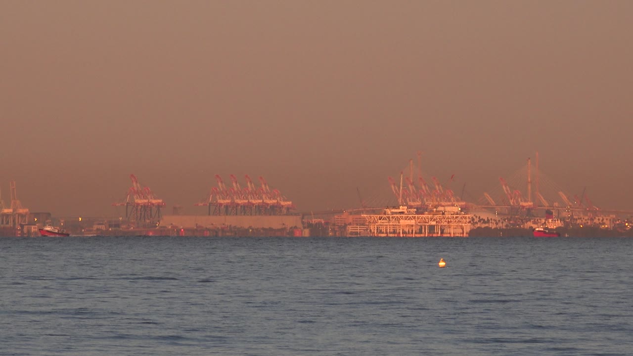 Small boat near distant cranes beyond a broad band of ocean water.