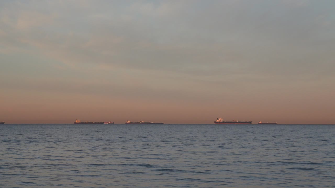 Wide dusk view of the Huntington Beach ocean with rippled water in front, several distant ships along the horizon, muted clouds above, and no visible people.
