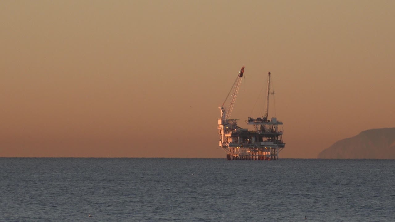 A distant offshore platform or large boat-like structure sits on the Huntington Beach ocean horizon under warm orange light, with no visible people.