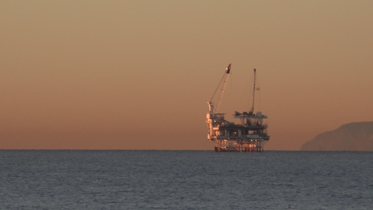 The Huntington Beach ocean fills the foreground while a crane-topped offshore structure remains near the horizon in warm dusk color, with no visible people.