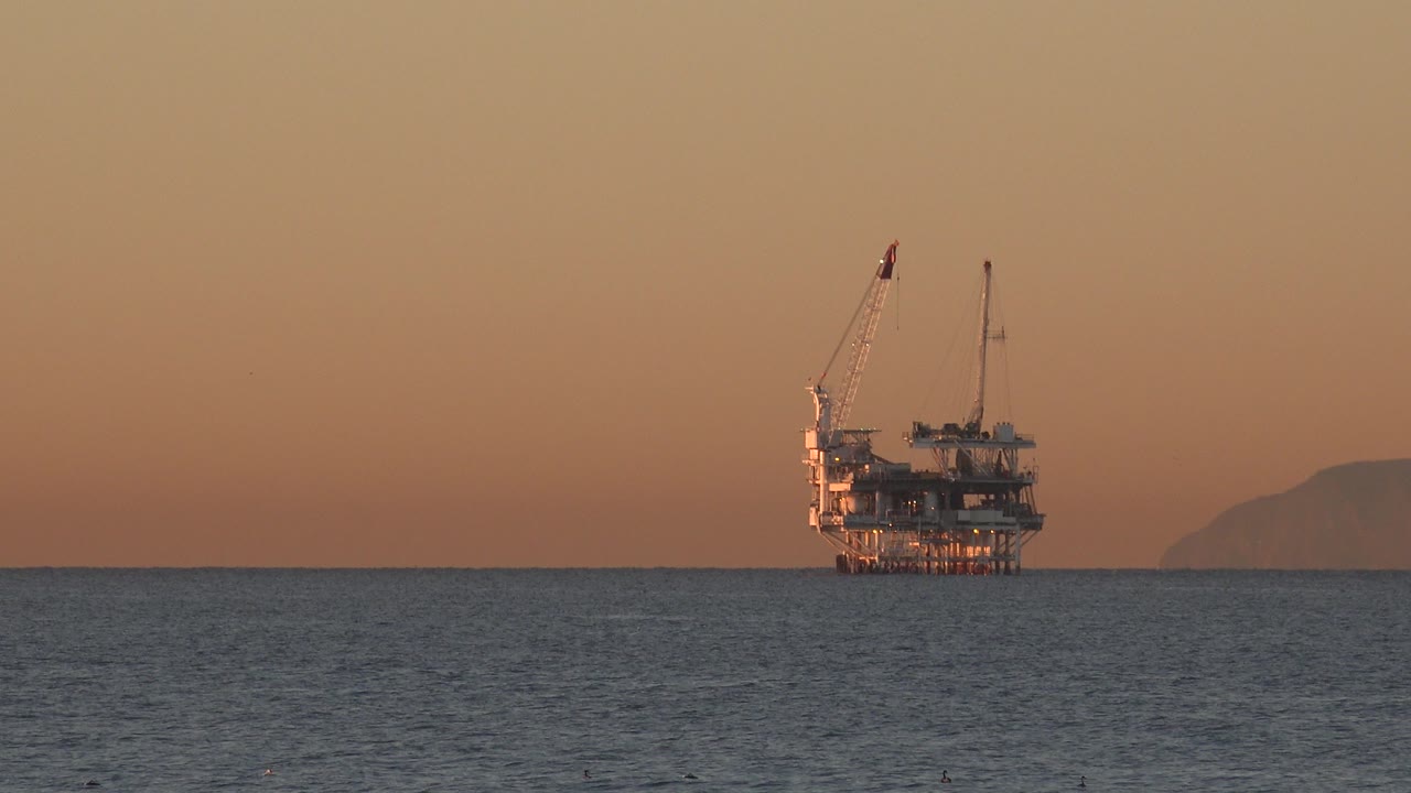 At 0:01, the offshore rig remains near the horizon at Huntington Beach, California, with rippled water in the foreground and no visible people.