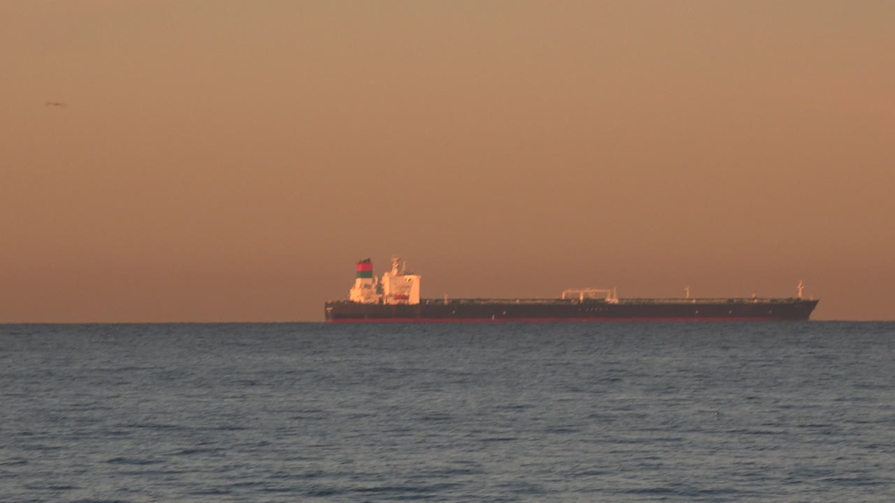 The ship appears near the center of the Huntington Beach horizon, its dark hull set against an orange sky, with textured ocean water below and no visible people.