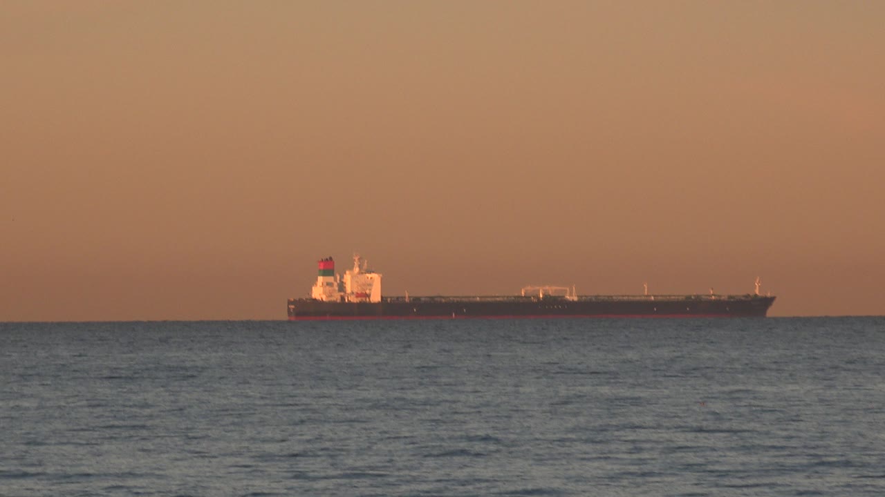 Large dark-hulled ship offshore at Huntington Beach, California, with calm water in the foreground, an orange sky above, and no visible people.