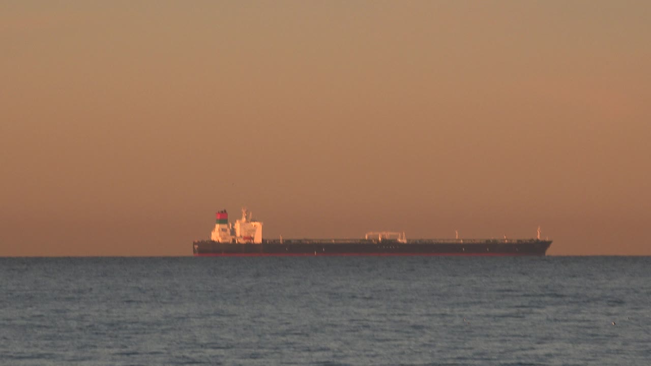 A distant tanker ship rests along the horizon off Huntington Beach, California, with rippled ocean foreground, orange sunset sky, and no visible people.