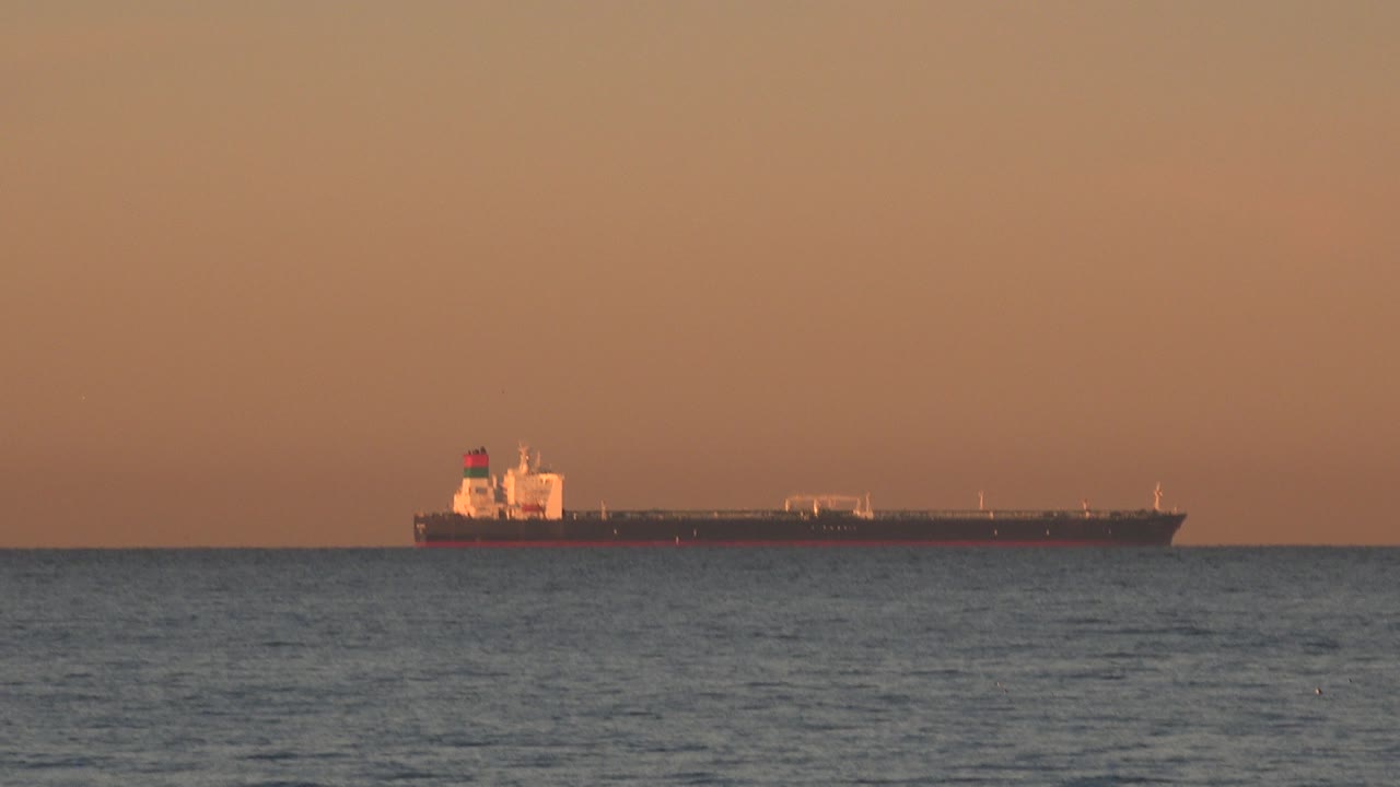 The tanker remains near the ocean horizon off Huntington Beach, California, with the bow and superstructure catching warm light and no visible people.
