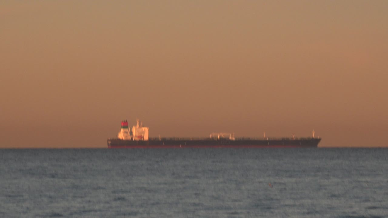 Long tanker ship silhouetted against orange sky over open water.