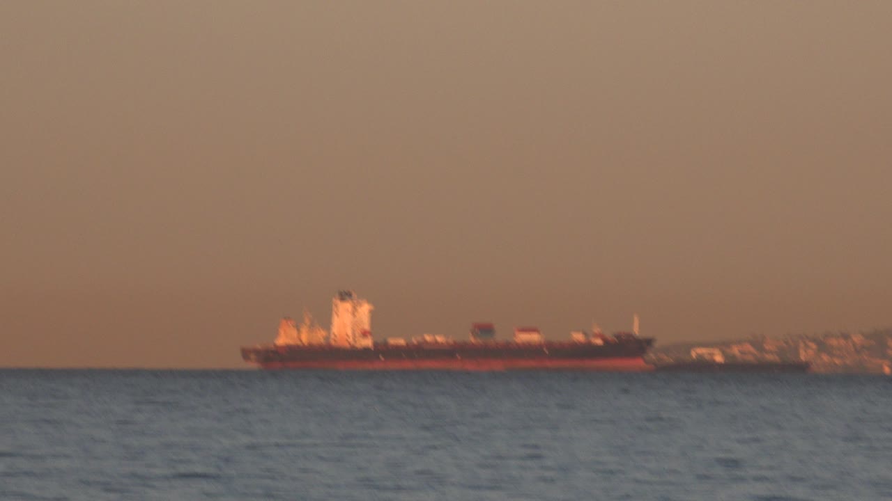 A distant oil tanker sits offshore near Huntington Beach, California, with rippled water in the foreground, low shoreline structures behind it, and no visible people.