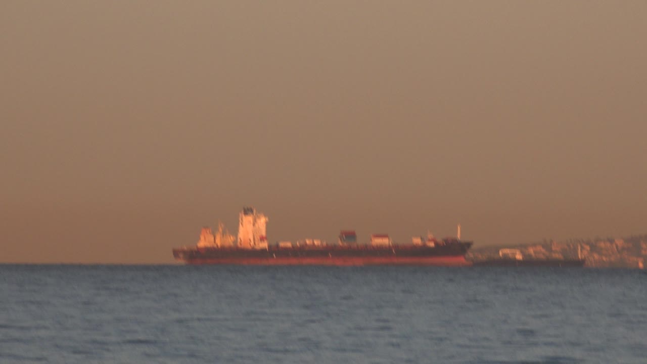 The tanker remains centered offshore at Huntington Beach, California, with small surface waves below it, a hazy industrial shoreline behind, and no visible people.