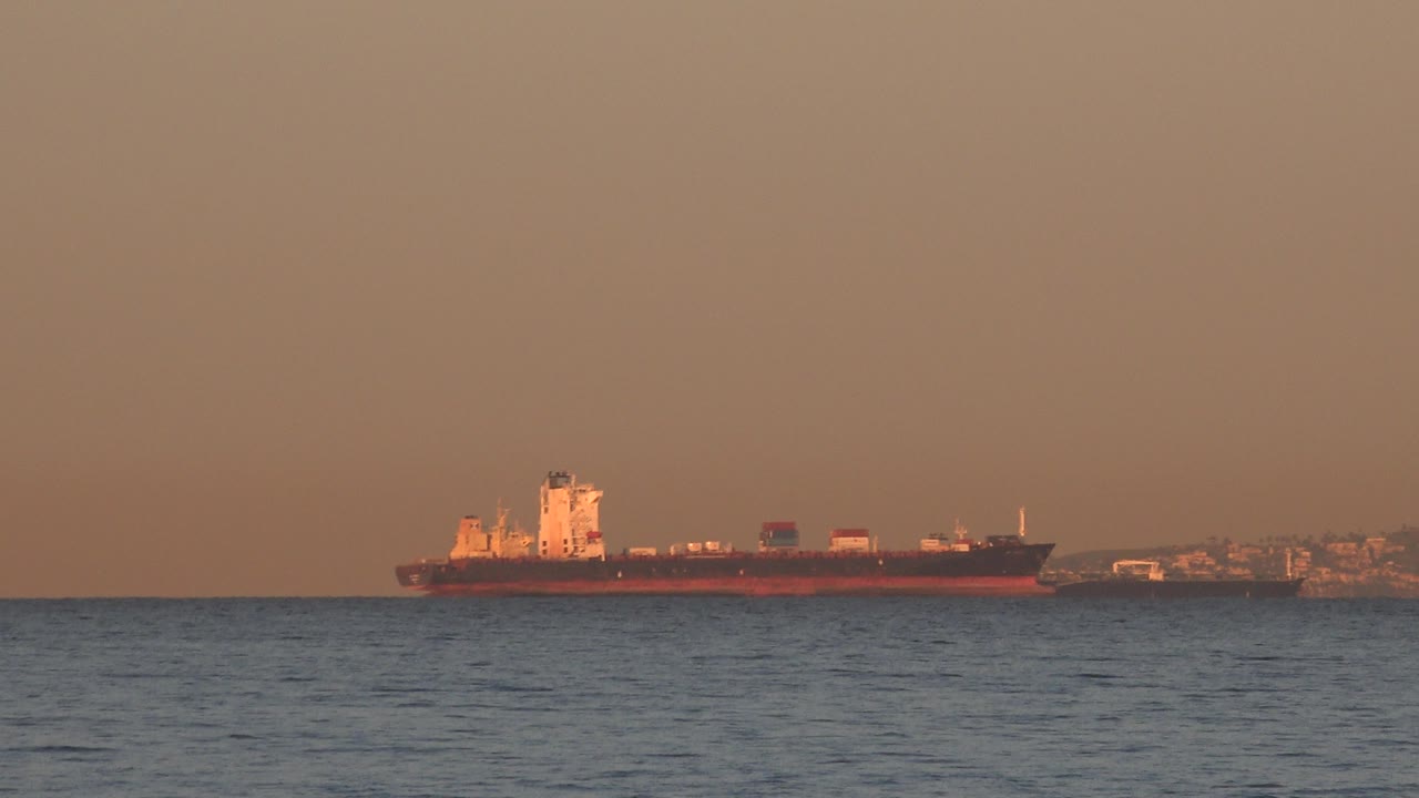 At 0:00, a large cargo ship sits in the distance off Huntington Beach, California, with blue ocean in the foreground, a hazy orange sky, shoreline behind it, and no visible people.