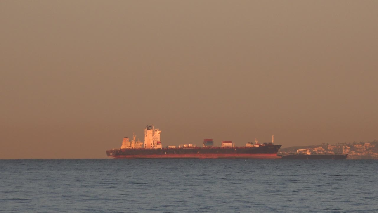 At 0:01, the container ship remains centered across the water near Huntington Beach, California, with low port structures and shoreline visible behind it and no visible people.