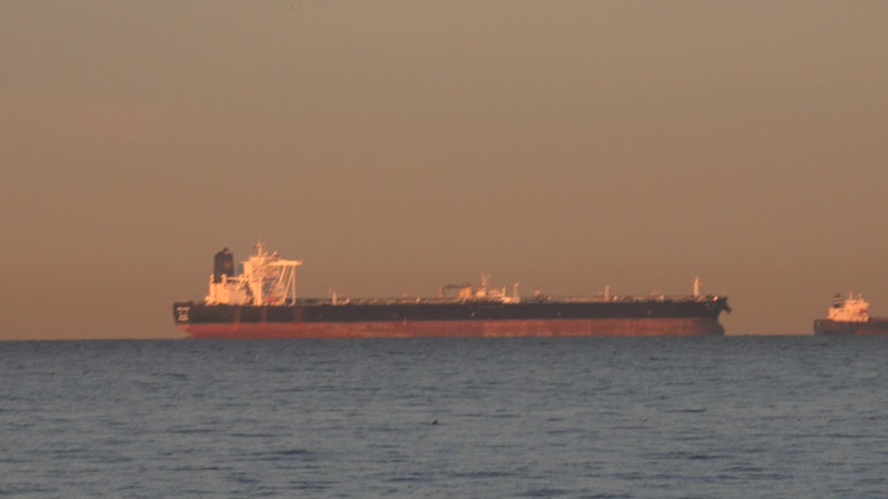 At 0:00, a large red and black boat floats far offshore at Huntington Beach, California, with a small boat to the right, blue water in front, and no visible people.