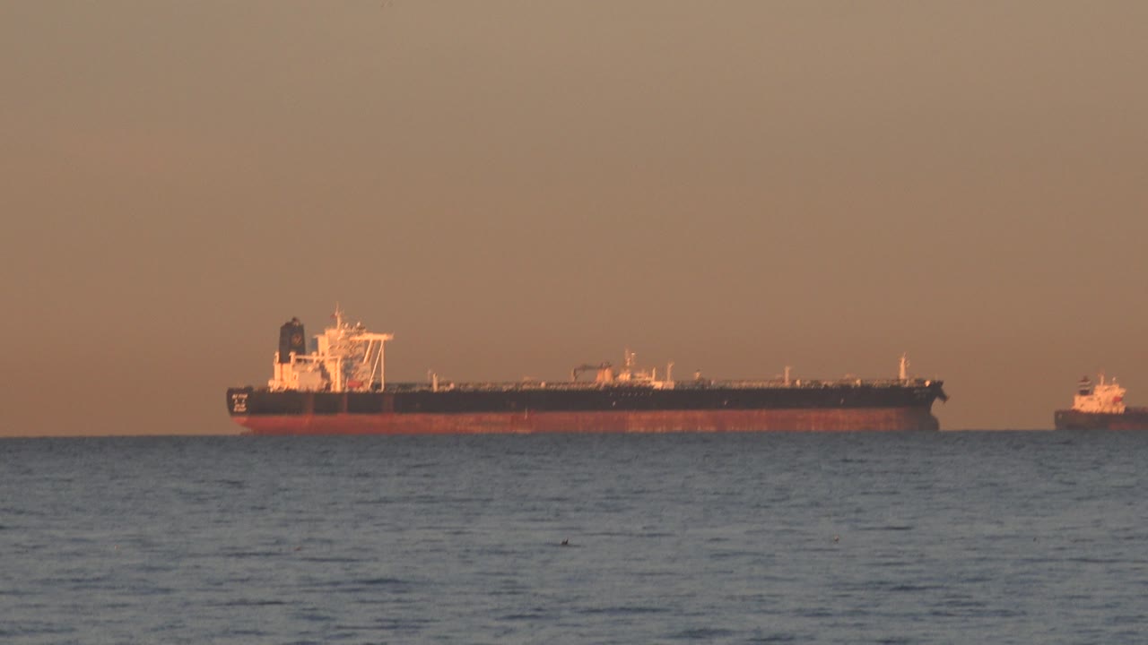 At 0:01, the distant vessel remains centered against the orange sky at Huntington Beach, California, with a small boat separated to the right and no visible people.
