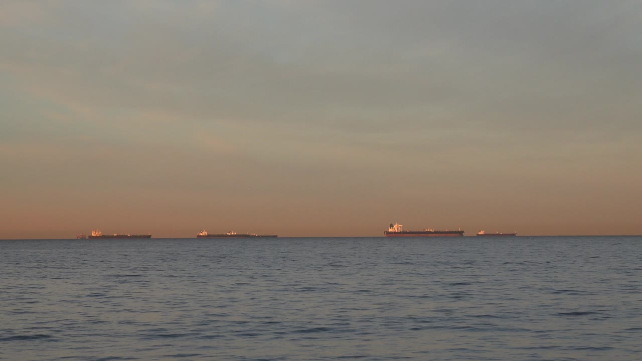 Huntington Beach seascape with several cargo-like vessels appearing as dark silhouettes on the far horizon, soft orange banding near the water, and no visible people.
