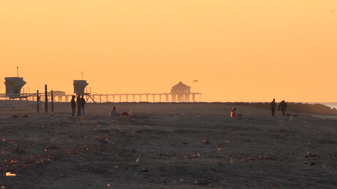 Huntington Beach at dusk with dark sand in the foreground, a warm orange sky, small birds overhead, distant lifeguard structures, and tiny silhouetted beachgoers near the horizon.