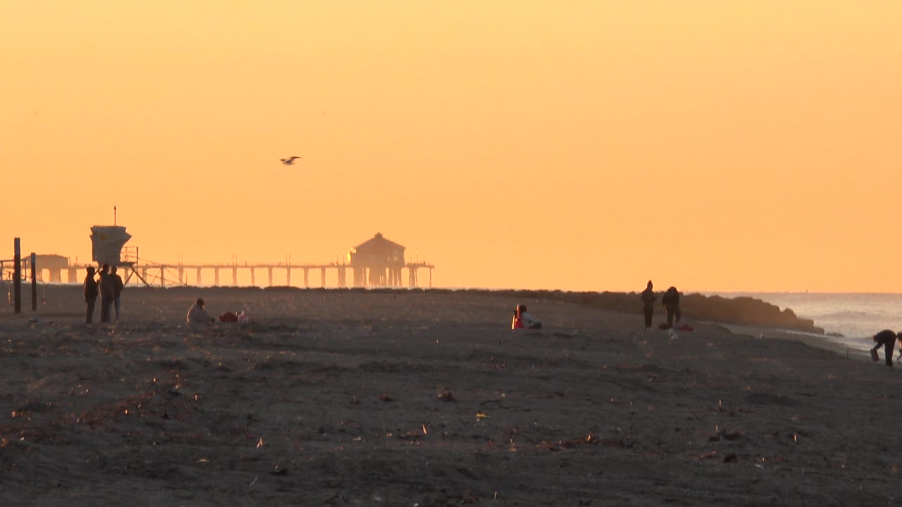 Huntington Beach wide shot at the start of the clip, with scattered silhouetted people on rough sand, a lifeguard tower at left, the pier in the distance, and an orange hazy sky.