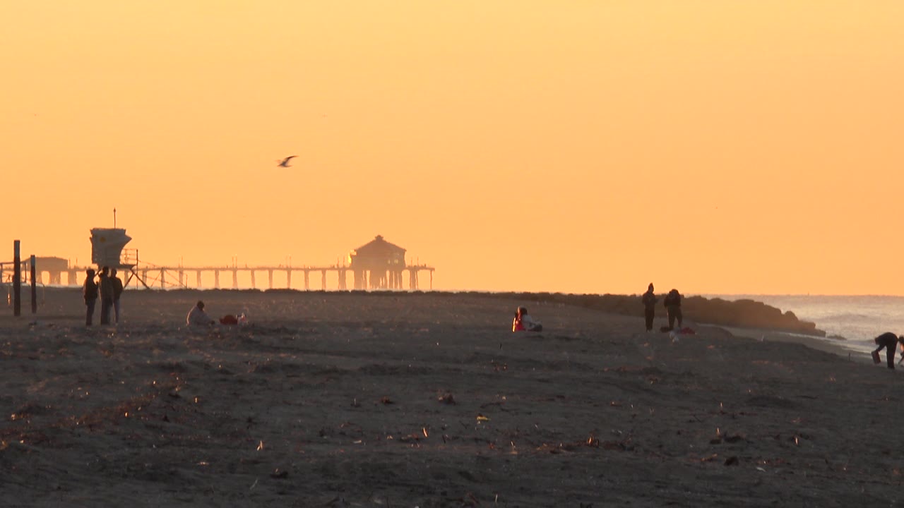 Huntington Beach frame with silhouetted beachgoers near the lifeguard tower, a person low to the sand near center, the pier crossing the background, and the ocean line at right.