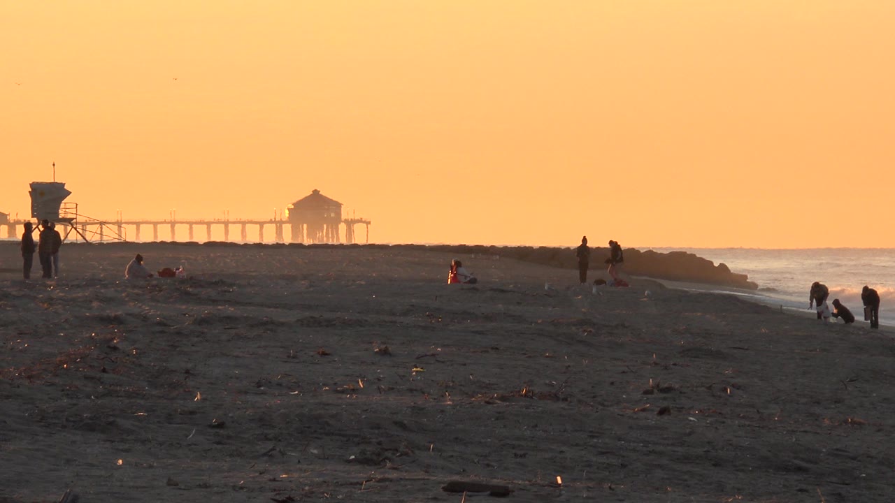 Sunset beach with distant pier, small people, and waves at right.