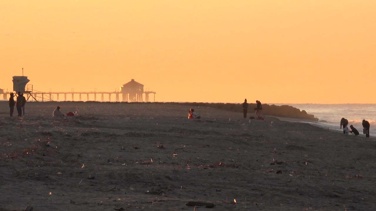 Huntington Beach scene with silhouetted beachgoers spread across dark sand, a red-jacketed child in the midground, surf along the right edge, and the pier in the distance.