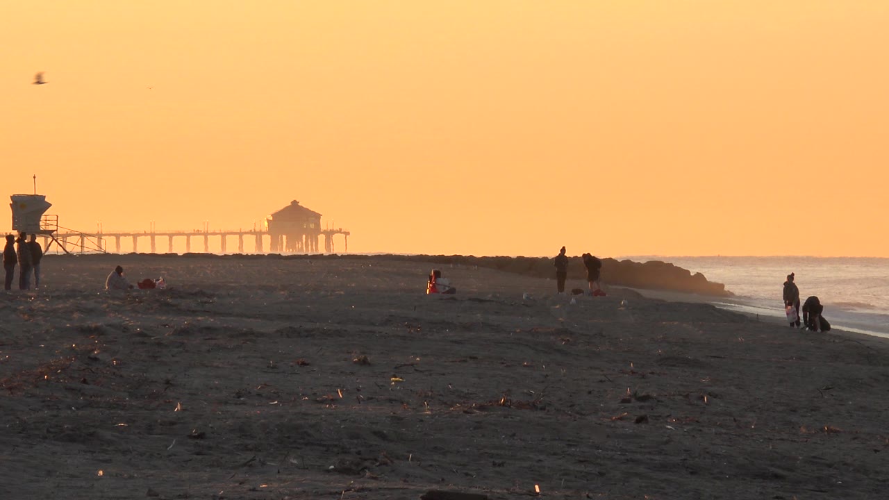 Huntington Beach sunset view with a dark sandy foreground, small distant people near the shoreline, a lifeguard tower at left, and the pier silhouetted on the horizon.