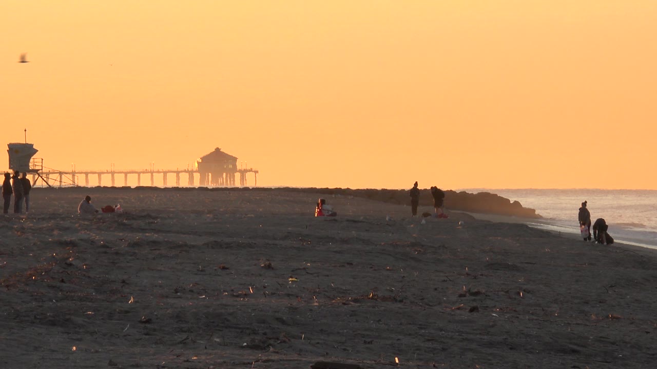 At 0:01, the Huntington Beach shoreline remains in orange light, with the lifeguard tower near center-left, the pier behind it, and small silhouetted beachgoers spread along the sand.