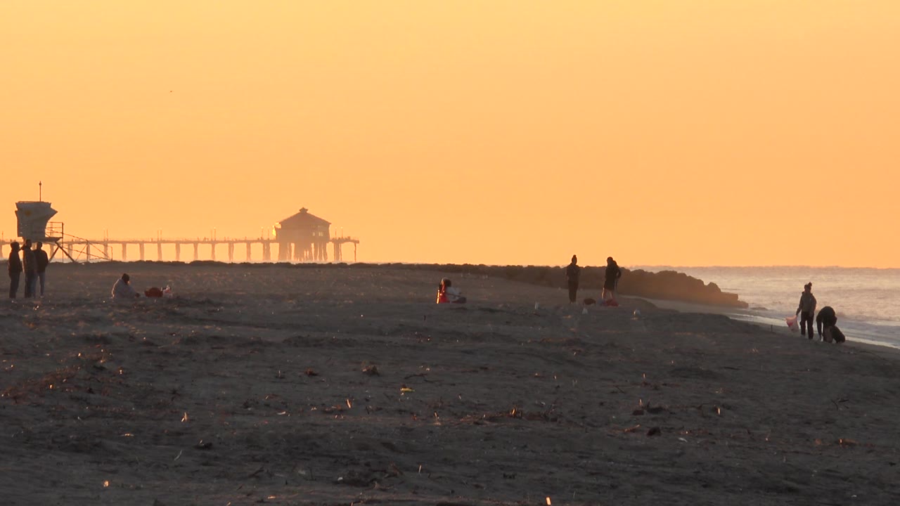At 0:02, the Huntington Beach pier sits left of center in the background while scattered distant silhouettes stand or crouch along the dark beach beside the ocean.