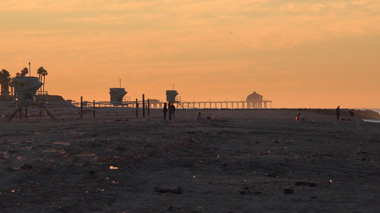 Sunset beach with lifeguard towers, distant people, and a pier silhouette.