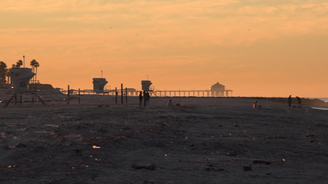 Huntington Beach, California beach at sunset with dark sand in front, lifeguard towers on the left, small distant people, and a pier silhouetted on the horizon.