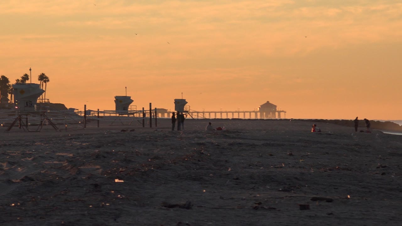 Huntington Beach, California shoreline with two silhouetted people near beach posts, lifeguard towers to the left, and the pier faintly lit by the orange sky.