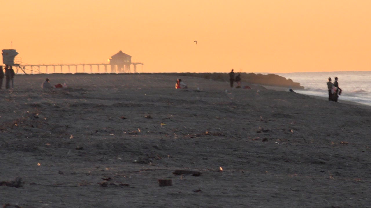 At 0:00, Huntington Beach appears in orange low light with dark sand in front, distant people near the shoreline, small birds in the sky, and a pier-like structure on the horizon.