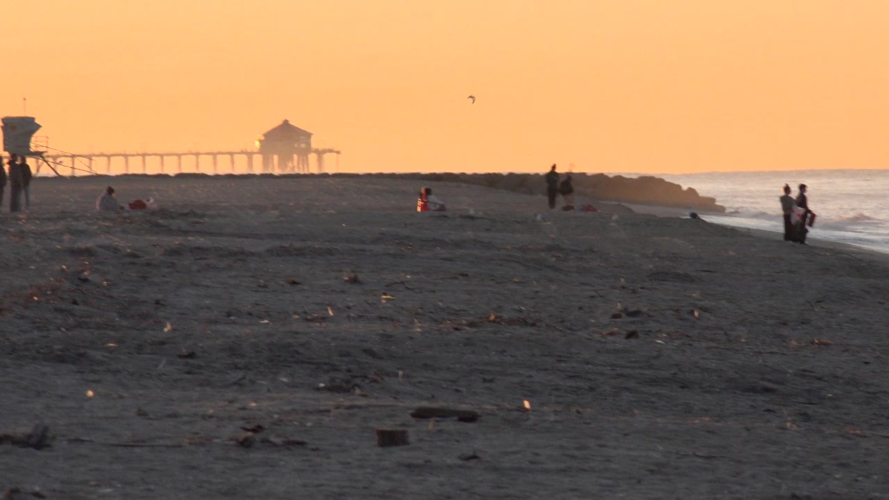 At 0:01, the Huntington Beach shoreline remains wide and low, with a few distant figures on the sand, small birds overhead, surf at right, and the horizon structure shifted slightly in frame.
