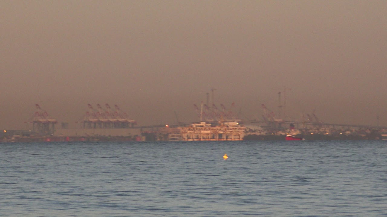 At 0:00, Huntington Beach ocean water fills the foreground with distant cargo ships, port cranes, small red boats, and hazy orange sky on the horizon, with no visible people.