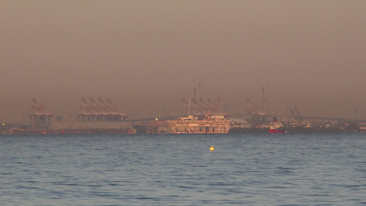 At 0:01, the same Huntington Beach shoreline view shows rippled blue water below distant cargo vessels and clustered cranes under warm haze, with no visible people.